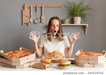 Photo of amazed excited woman with brown hair wearing white T-shirt sitting at table with raised arms and open mouth, being ready to eat pasta, hamburger, pizza and other junk food. 104329742