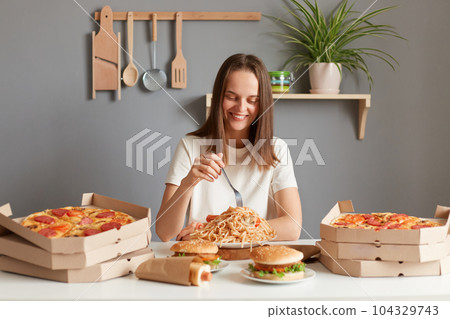 Portrait of charming cute pretty woman with brown hair wearing white T-shirt sitting at table eating pasta, holding fork in hand, enjoying her dinner with fast food. 104329743