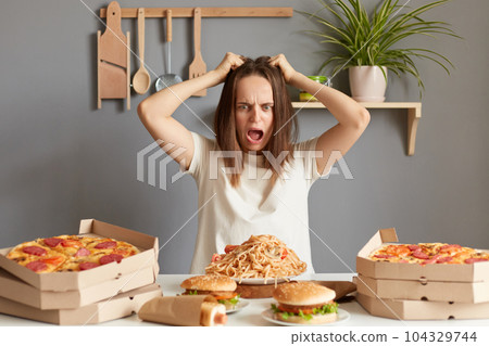 Portrait of crazy irritated shocked woman wearing white T-shirt sitting at table in kitchen, pulling her hair, a girl on a diet, surrounded by delicious junk food, does not know how not to break loose 104329744