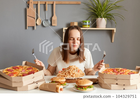 Horizontal shot of satisfied woman with brown hair wearing white casual T-shirt sitting at table in kitchen, posing with closed eyes, smelling pasta, pizza, hamburgers. Horizontal shot of satisfied woman with brown hair wearing white casual T-shirt sitting at table in kitchen, posing with closed eyes, smelling pasta, pizza, hamburgers. 104329751