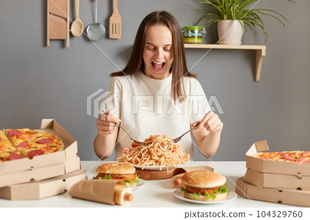 Portrait if amazed excited extremely hungry woman wearing white T-shirt sitting at table in kitchen, delivering lots jink food dishes, eating noodles, expressing happiness. 104329760