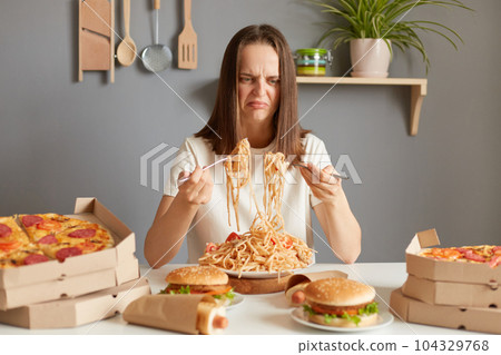 Portrait of disappointed sad woman wearing white T-shirt sitting at table in kitchen, looking at pasta, thinks she delivering spoiled food, frowning face. 104329768