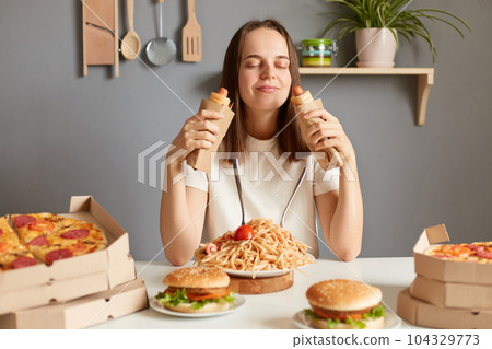 Image of satisfied cheerful young adult Caucasian woman wearing white casual T-shirt sitting at table in kitchen, holding in hands two hot dogs, smelling delicious aromat with closed eyes. 104329773