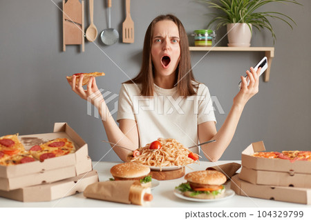 Indoor shot of shocked irritated woman with brown hair wearing white t shirt sitting in kitchen at table and holding smart phone and slice pizza, looking at camera with shocked face. Indoor shot of shocked irritated woman with brown hair wearing white t shirt sitting in kitchen at table and holding smart phone and slice pizza, looking at camera with shocked face. 104329799