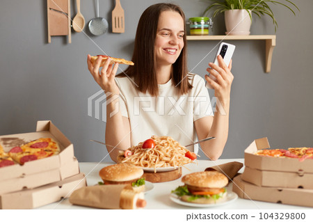 Portrait of attractive charming woman with brown hair sitting in kitchen and using mobile phone while eating tasty pizza, chatting with friends, left positive feedback delivery service. 104329800
