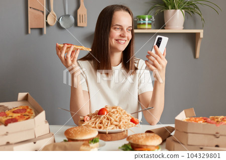 Photo of cheerful Caucasian young adult woman with brown hair wearing white t shirt sitting in kitchen at table and looking at smart phone display, having delicious snack, eating pizza. 104329801