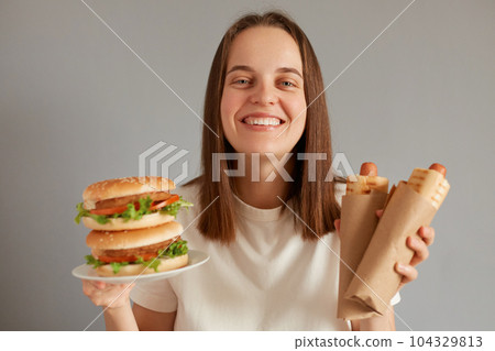 Portrait of happy smiling woman holding hot dog and burger wearing white T-shirt posing isolated over gray background, looking at camera with toothy smile, enjoying eating fast food. 104329813