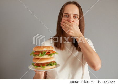 Indoor shot of unhealthy woman feeling sick, covering her mouth after eating burger, young person suffering indigestion feeling the need to vomit after a spoiled sandwich. Indoor shot of unhealthy woman feeling sick, covering her mouth after eating burger, young person suffering indigestion feeling the need to vomit after a spoiled sandwich. 104329826