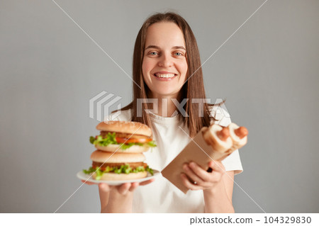 Horizontal shot of smiling satisfied female with brown hair, standing isolated on gray background, holding hot dogs and american hamburger, enjoying fast food. Horizontal shot of smiling satisfied female with brown hair, standing isolated on gray background, holding hot dogs and american hamburger, enjoying fast food. 104329830