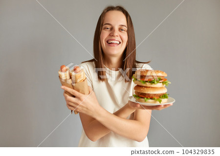 Portrait of laughing woman holding tasty cheeseburger and hotdogs, crossed hands, looking at camera with cheerful happy expression, likes unhealthy food. 104329835