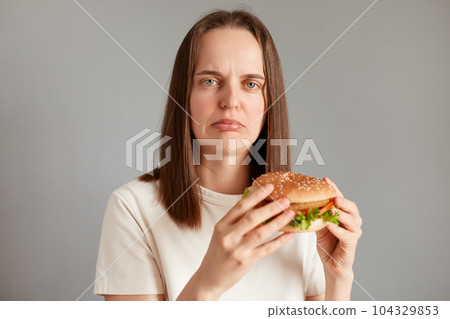 Indoor shot of sad attractive woman keeps diet, feels hungry, holding american hamburger in hands, looking at camera with sorrow, needs to keep healthy eating. 104329853