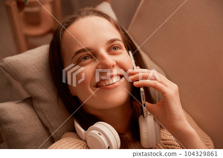Top view of smiling happy young caucasian girl holding talking via mobile phone, expressing positive emotions, laying on sofa at home, having pleasant conversation. 104329861