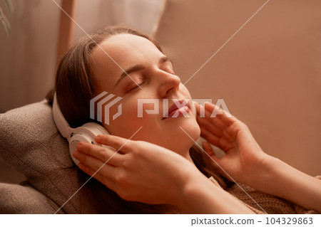 Side view portrait of relaxed young adult woman listening music while resting on couch at home, keeps eyes closed, touching her headphones, enjoying moment. 104329863