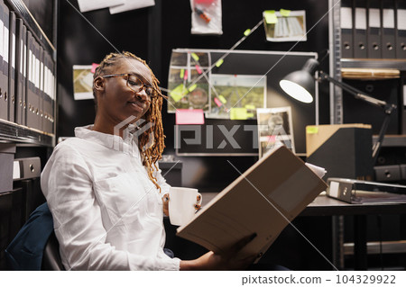 Policewoman reading investigation report while drinking coffee in office. African american woman police detective holding crime case file folder and analyzing evidence at night Policewoman reading investigation report while drinking coffee in office. African american woman police detective holding crime case file folder and analyzing evidence at night 104329922