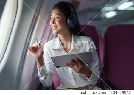 Young Asian attractive business woman passenger sitting on business class luxury plane while wearing wireless headphone for relax and working with tablet during the flight. Young Asian attractive business woman passenger sitting on business class luxury plane while wearing wireless headphone for relax and working with tablet during the flight. 104329947