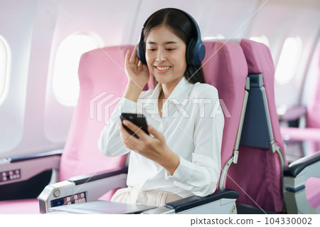 Young Asian attractive business woman passenger sitting on business class luxury plane while wearing wireless headphone and using smart phone mobile for relax during the flight. Young Asian attractive business woman passenger sitting on business class luxury plane while wearing wireless headphone and using smart phone mobile for relax during the flight. 104330002