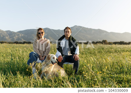 Young happy couple with dog, sitting on chairs in feather grass field on summer sunset day, looking forward in camera, serious Young happy couple with dog, sitting on chairs in feather grass field on summer sunset day, looking forward in camera, serious 104330009
