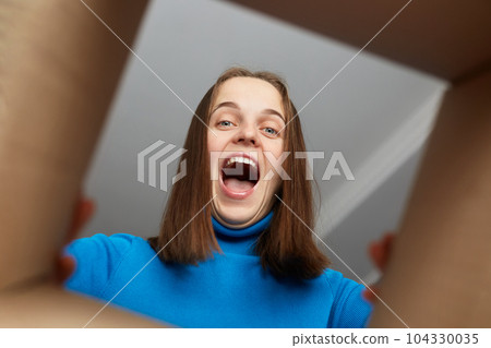Young adult Caucasian woman with brown hair unpacking carton box and trying to get something, looking inside with joyful expression, saying wow, view from the inside. 104330035
