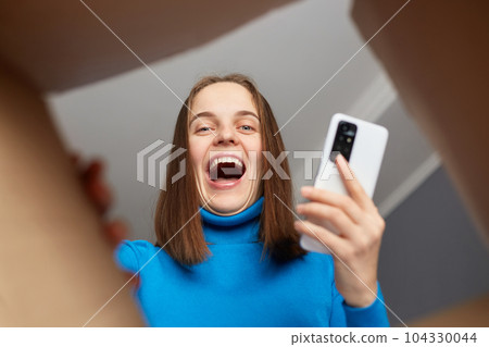 Extremely happy Caucasian woman unpacking box and looking in cardboard box with happy smile, looks joyful, holding smart phone in hands, received long awaited parcel, view from the inside of package. Extremely happy Caucasian woman unpacking box and looking in cardboard box with happy smile, looks joyful, holding smart phone in hands, received long awaited parcel, view from the inside of package. 104330044
