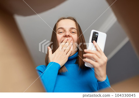 Positive optimistic Caucasian woman customer wearing blue turtleneck opening carton box, looking inside package with happy surprised face, covering mouth, using mobile phone. Positive optimistic Caucasian woman customer wearing blue turtleneck opening carton box, looking inside package with happy surprised face, covering mouth, using mobile phone. 104330045