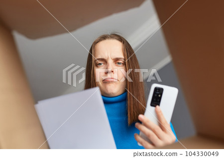 Sad serious woman customer wearing blue turtleneck opening parcel, looking at consignment, trying to understand where is a mistake, holding phone, needs to call to delivery service. Sad serious woman customer wearing blue turtleneck opening parcel, looking at consignment, trying to understand where is a mistake, holding phone, needs to call to delivery service. 104330049