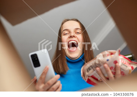 Happy excited joyful woman receives a package from online shop order, female with brown hair has glad expression, taking of products and holding cell phone, being satisfied, view from the box. Happy excited joyful woman receives a package from online shop order, female with brown hair has glad expression, taking of products and holding cell phone, being satisfied, view from the box. 104330076