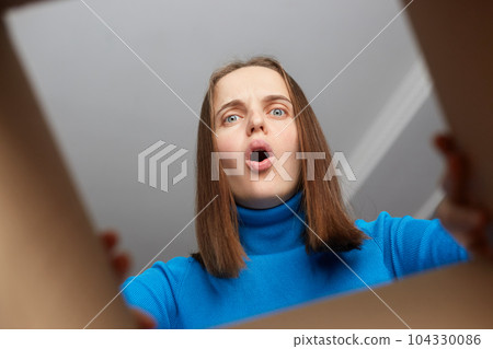 Shocked disappointed woman customer wearing blue turtleneck opening cardboard box with broken or wrong order, looking inside parcel with widely open mouth and big eyes. Shocked disappointed woman customer wearing blue turtleneck opening cardboard box with broken or wrong order, looking inside parcel with widely open mouth and big eyes. 104330086