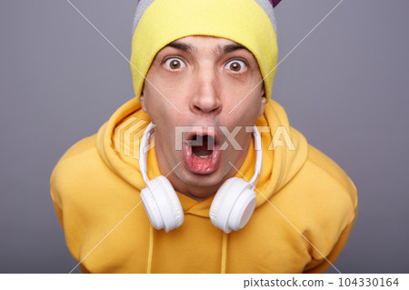 Closeup portrait of amazed surprised astonished hipster man wearing yellow hoodie and beanie hat standing against grey wall expressing shock, looking at camera with big eyes and open mouth. 104330164