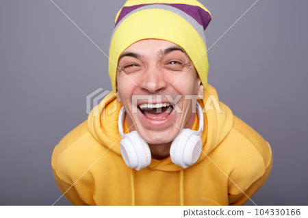 Closeup portrait of extremely happy cheerful hipster man wearing yellow hoodie and beanie hat, hearing funny joke or watching video, laughing out loud, standing against grey wall expressing 104330166