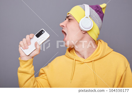 Image of funny cheerful young adult man in beanie hat and yellow casual hoodie, listening his favorite playlist, holding smart phone as a microphone, singing, standing isolated over gray background. 104330177