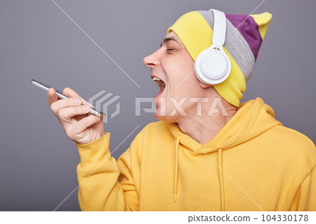 Horizontal shot of attractive delighted cheerful man in yellow hoodie and wireless headphones, singing songs, holding smartphone, recording message with his song, posing isolated over grey background. 104330178