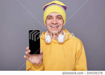 Smiling kind friendly man in beanie hat and yellow casual hoodie, holding mobile phone with blank screen, standing isolated over gray background, showing phone with empty display for advertisement. 104330179