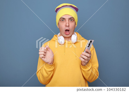Horizontal shot of shocked amazed man wearing beanie hat and yellow hoodie, holding smart phone in hands, reading breaking new in internet, looking at camera with open mouth, posing against blue wall 104330182