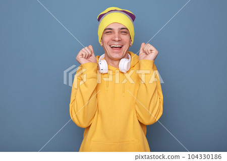 Portrait of extremely happy overjoyed man wearing beanie hat and casual yellow hoodie, raised his clenched fists, rejoicing his success, celebrating, smiling happily, posing against blue wall Portrait of extremely happy overjoyed man wearing beanie hat and casual yellow hoodie, raised his clenched fists, rejoicing his success, celebrating, smiling happily, posing against blue wall 104330186