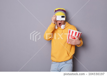 Indoor shot of astonished anonymous man dressed in yellow hoodie, jeans and beanie hat, visiting cinema, poses against gray wall, holding paper bucket with popcorn, covering his eyes with cell phone. 104330197