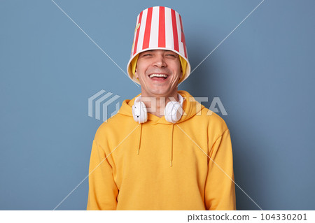 Portrait of happy overjoyed man wearing beanie hat and casual yellow hoodie posing against blue wall, standing with popcorn bucket on head, laughing out loud, having fun in cinema 104330201