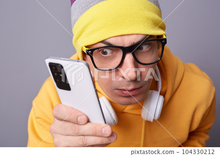 Closeup shot of funny attentive man gazes with surprised expression at mobile phone display wears casual closing isolated over gray background looks with big interest at smart phone. 104330212