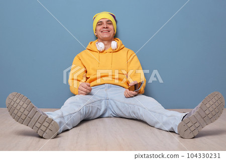 Horizontal shot of smiling relaxed man wearing beanie hat and casual yellow hoodie standing isolated over blue background, sitting on floor, looking at camera, resting, holding phone in hand 104330231