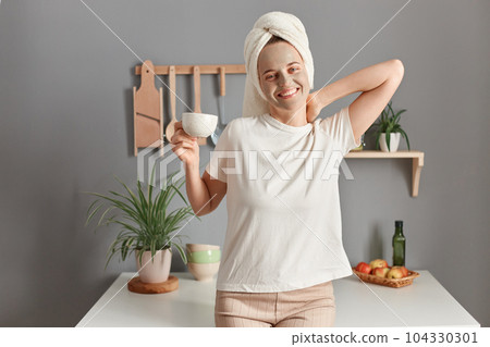 Indoor shot of smiling attractive positive woman with cosmetic mask on her face drinking coffee on the kitchen at home, wakes up in good mood, doing morning procedure for skin care. 104330301