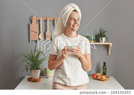 Smiling dreaming joyful woman with cleansing mask on her face drinking coffee on the kitchen at home, enjoying lazy morning, looking away with happy expression, enjoys hot beverage. 104330303