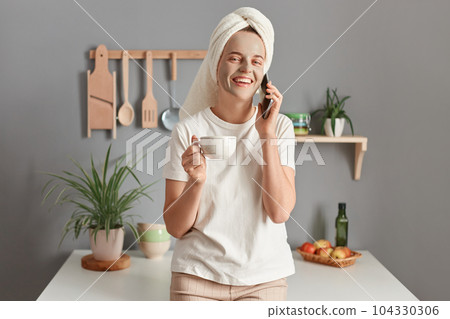 Joyful attractive young adult woman with cosmetic mask on her face drinking coffee on the kitchen at home, expressing positive emotions, talking on smart phone. Joyful attractive young adult woman with cosmetic mask on her face drinking coffee on the kitchen at home, expressing positive emotions, talking on smart phone. 104330306