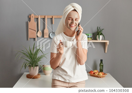Pretty woman with cosmetic mask on face drinking morning coffee and talking via smartphone at home in kitchen, young female wrapped in towel answering call by phone during beauty skincare treatment. 104330308