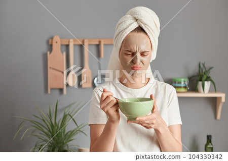 Sad upset woman wearing white t-shirt and wrapped towel on her hair doing cosmetic procedures and eating awful breakfast, thinking fie, frowning face from disgust. 104330342