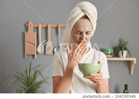 Upset Caucasian woman wearing white t-shirt and wrapped towel on her hair doing cosmetic procedures and having breakfast, covering mouth with palm, realized that the food is spoiled 104330346