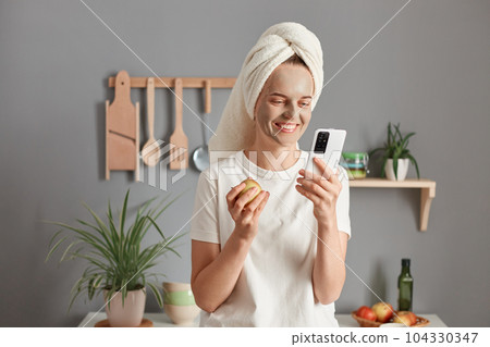 Indoor shot of smiling woman with treatment facial mask enjoying spa procedures at home, posing in the kitchen, eating apple and using cell phone, checking social networks. Indoor shot of smiling woman with treatment facial mask enjoying spa procedures at home, posing in the kitchen, eating apple and using cell phone, checking social networks. 104330347