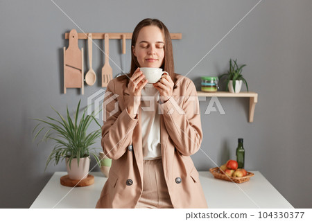Horizontal shot of joyful young adult woman wearing beige suit standing near table on kitchen at home, holding cup of coffee, keeps eyes closed, enjoying tasty smell of coffee. Horizontal shot of joyful young adult woman wearing beige suit standing near table on kitchen at home, holding cup of coffee, keeps eyes closed, enjoying tasty smell of coffee. 104330377