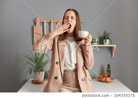 Portrait of sleepy brown haired woman wearing beige suit standing near table on kitchen at home, drinking coffee to have energy after early wake up, yawning, covering mouth with hand. 104330378