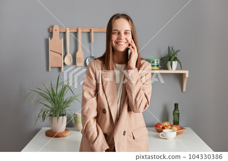 Portrait of cheerful joyful brown haired woman wearing beige suit standing near table on kitchen at home, looking at camera, talking phone with her friend. Portrait of cheerful joyful brown haired woman wearing beige suit standing near table on kitchen at home, looking at camera, talking phone with her friend. 104330386