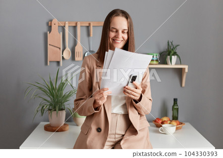 Image of attractive positive brown haired woman wearing beige suit standing near table on kitchen at home, holding smart phone, checking information in documents, enjoying her work. Image of attractive positive brown haired woman wearing beige suit standing near table on kitchen at home, holding smart phone, checking information in documents, enjoying her work. 104330393