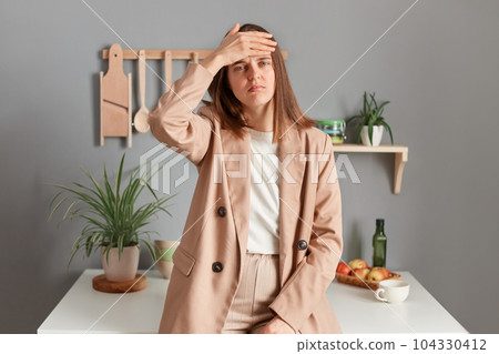 Portrait of sad sick ill young adult woman wearing beige suit standing near table on kitchen at home, keeps hand on forehead, suffering headache, having high temperature. 104330412